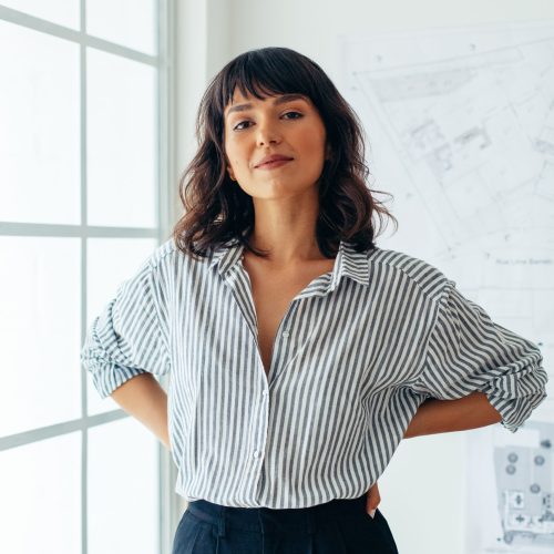 Portrait of businesswoman standing beside the window in office. Female architect standing in office with architecture drawings in the background.