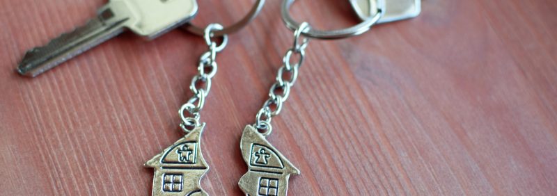 Two keys with splitted or broken key rings with pendant in shape of house divided in two parts on wooden background with copy space. Dividing house when divorce, division of property, real estate heri