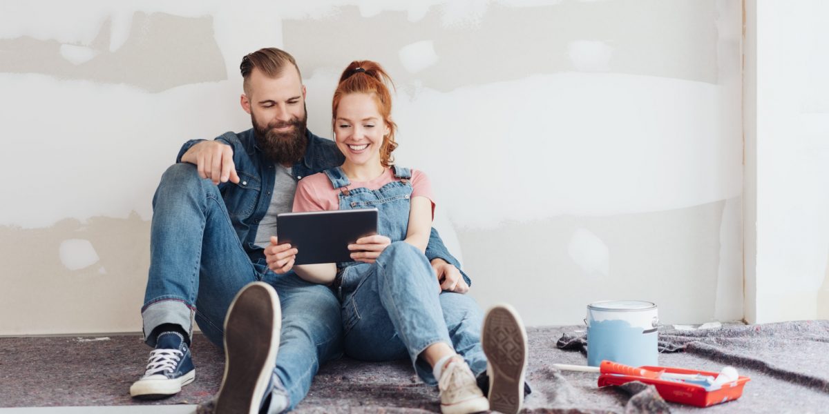 Young couple doing home renovations taking a break sitting on the floor leaning on an unfinished wall looking at a tablet pc with a smile